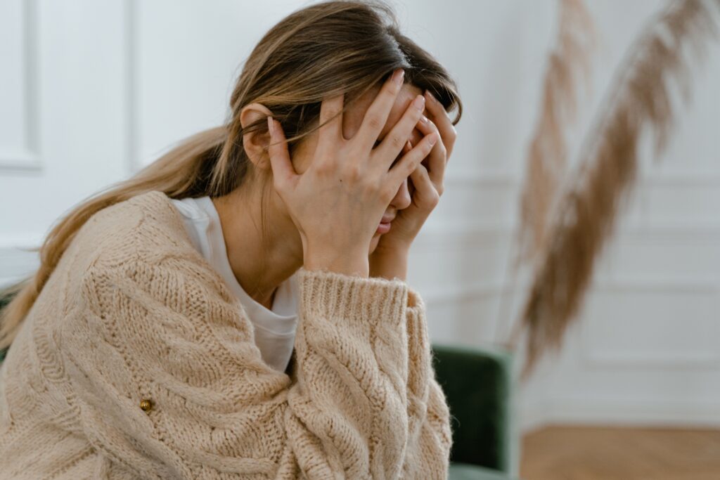 Os 5 Pensamentos Automáticos Mais Comuns na Ansiedade (e como identificar) A woman sitting indoors covering her face in frustration, depicting stress and mental health challenges.
